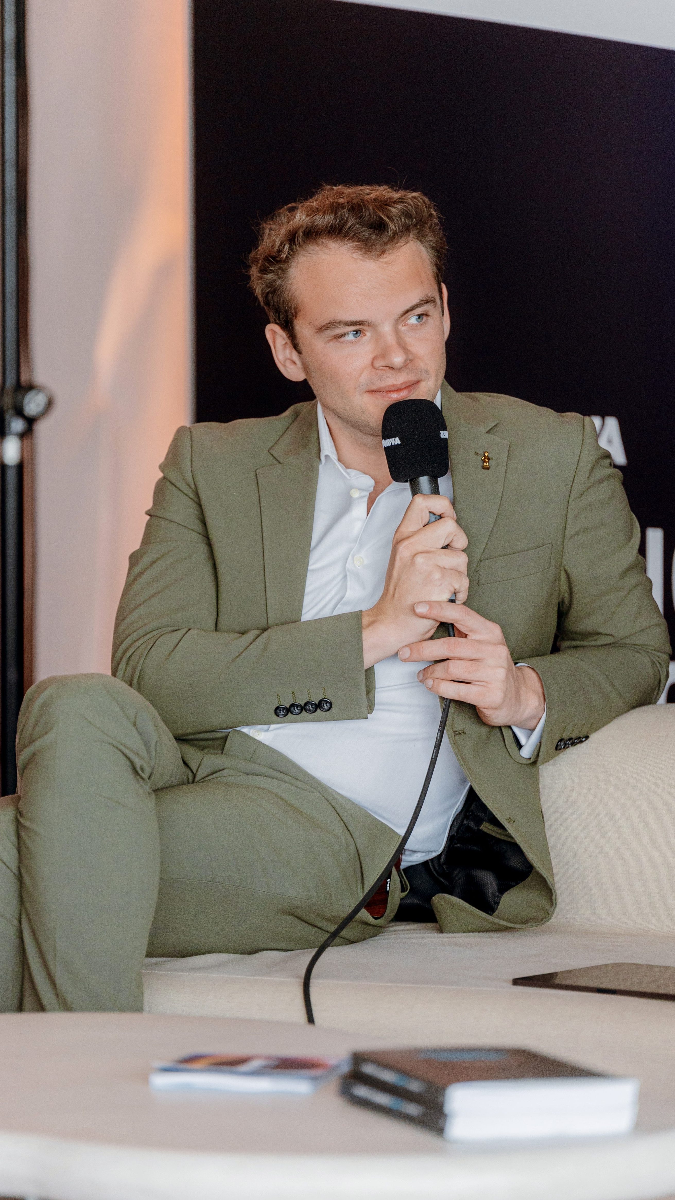 Professional host Wout, in an olive suit, listens intently while moderating a live panel, microphone in hand.